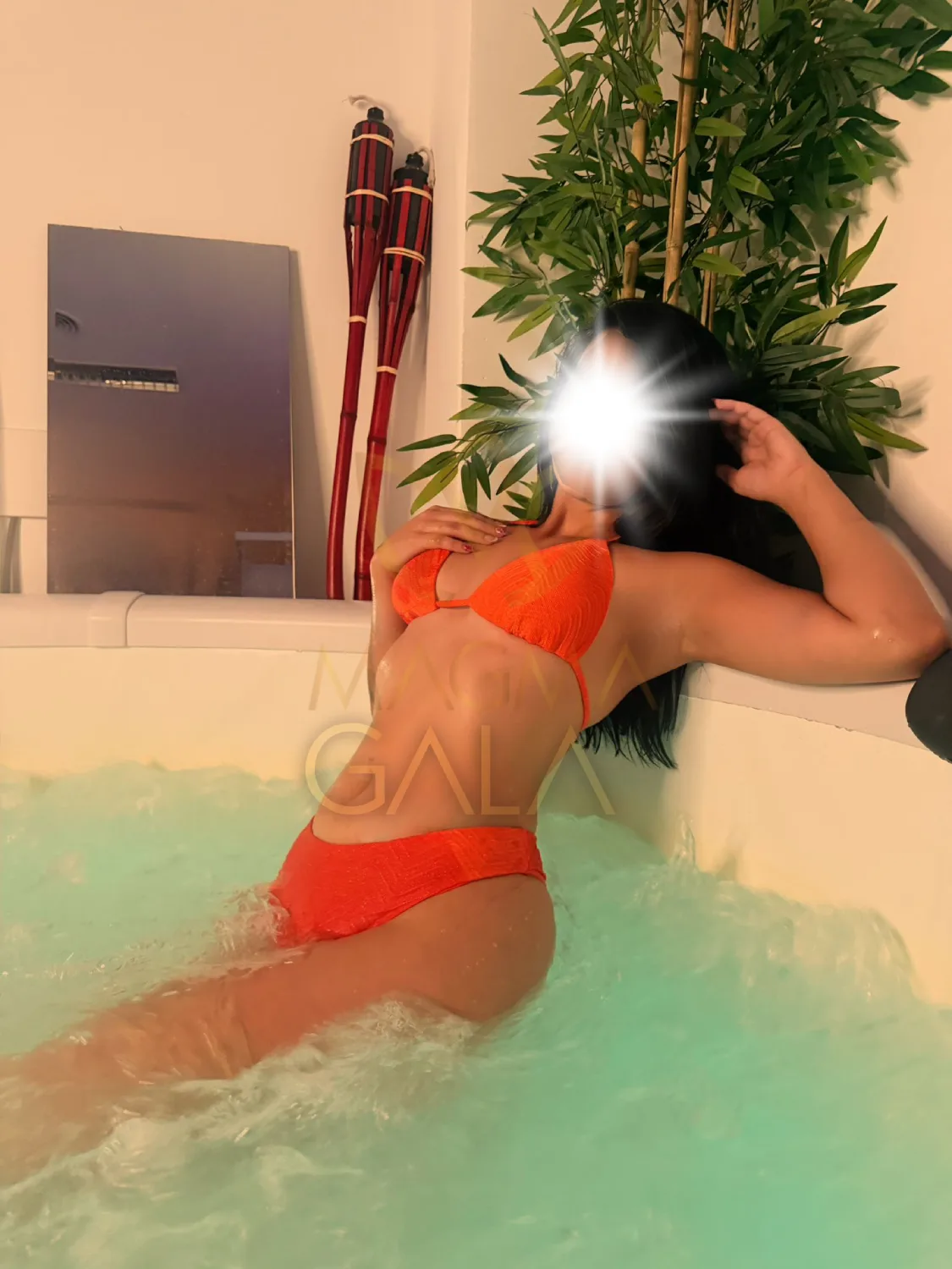 Gala in an orange bikini in the green-lit whirlpool bath, with one hand in her hair.
