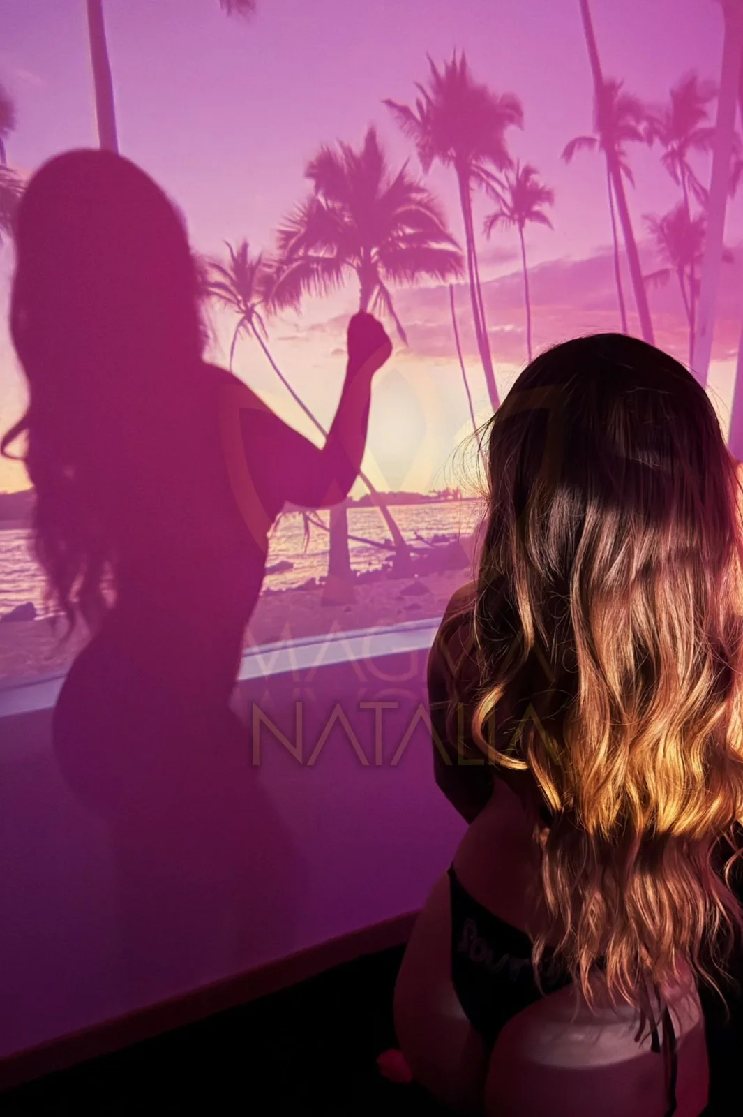 Natalia seen from behind, with long wavy hair, in front of a violet projection of palm trees.