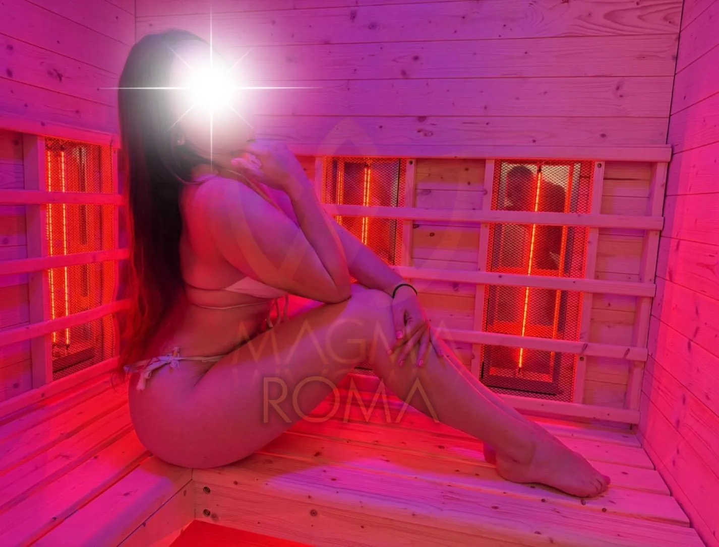 Roma in a pale outfit sitting in a wooden room lit in pink and violet.