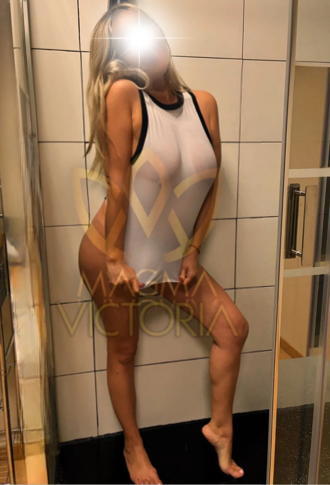 Victoria in a white swimsuit with black trim in a white-tiled shower.