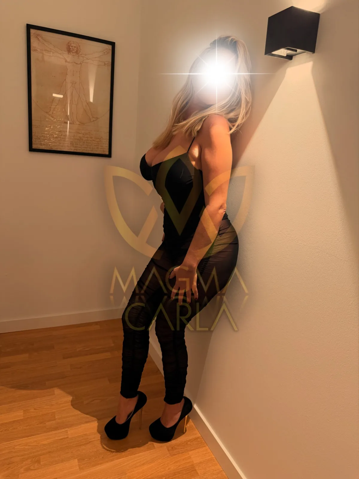 Victoria leaning against a wall in a white corridor, wearing a black outfit and heels.